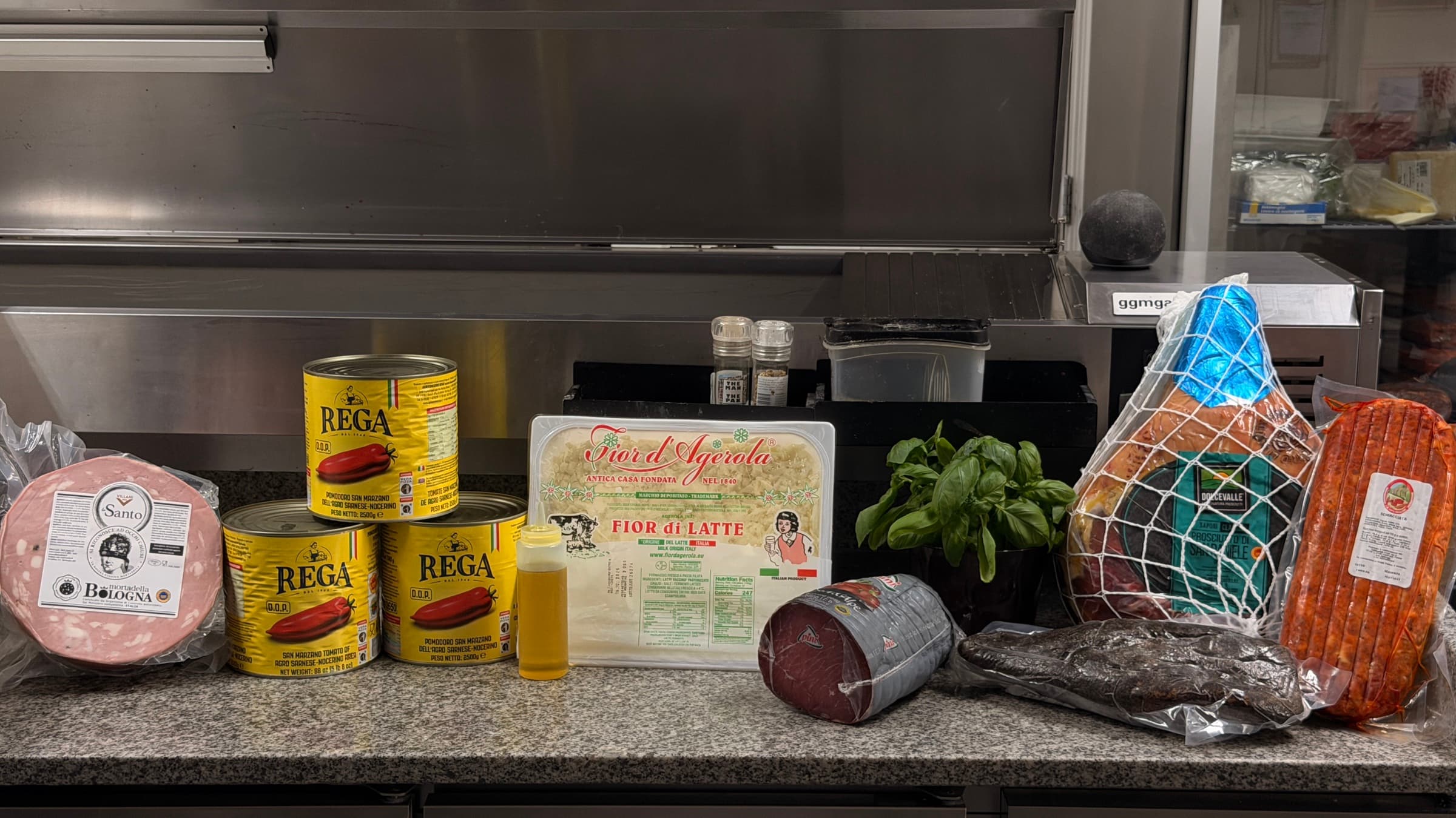 Italian ingredients on the prep counter: mortadella, tomato tins, fior di latte, basil, prosciutto and salami